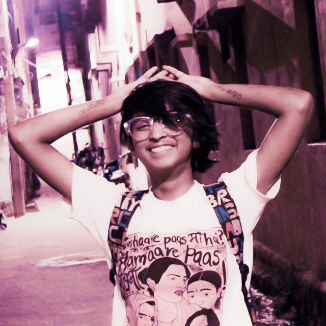 A young person with short black hair and glasses smiles widely while posing with hands raised in a narrow alleyway, wearing a t-shirt with a graphic design.